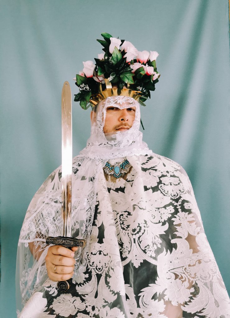 Artist Jose Campos holds sword in floral headdress, against turquoise curtain.